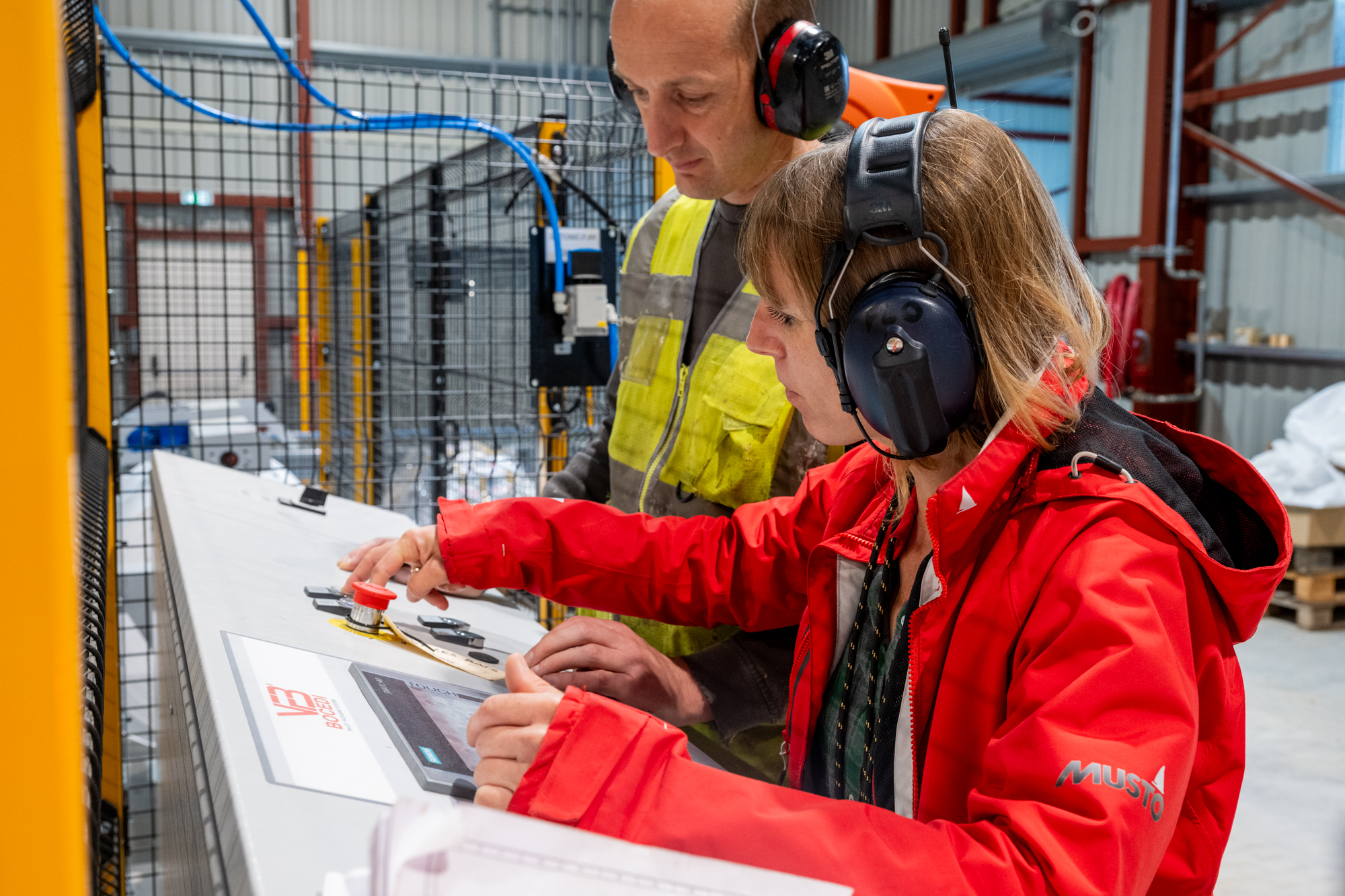 Deux opérateurs en tenue de sécurité effectuent un contrôle qualité sur une machine de production automatisée dans une usine de granulés de bois, portant des casques antibruit et EPI.